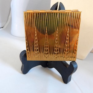 May include: A gold-colored, rectangular compact with a detailed Art Deco design. The compact features vertical lines and stylized wheat-like patterns. It is displayed on a small, black stand, showcasing its intricate details and vintage aesthetic.