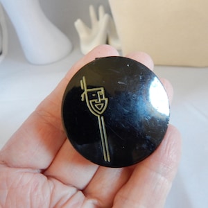 May include: A black, round compact with a gold-colored art deco design. The design features a stylized face with a long vertical line. The compact is held in a hand.