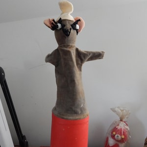 May include: A gray felt hand puppet mouse with black eyes and a white felt hat. The puppet is attached to a red cylinder.