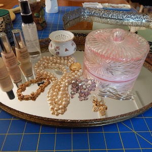 May include: An oval mirrored tray holds various items, including a clear, pink-lidded jar, pearl necklaces, a gold bracelet, and several bottles of makeup. A small, decorative floral container and gold earrings are also present.