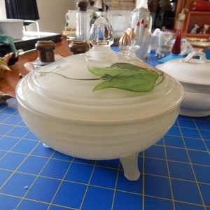 May include: A white glass lidded dish with a green leaf design on the lid. The dish has three feet and is sitting on a blue cutting mat.
