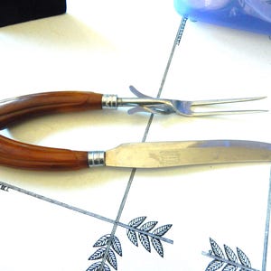 May include: A carving set featuring a stainless steel knife and fork. The knife has a long, curved blade and the fork has two tines. Both have brown handles with silver accents. The set is displayed on a white surface with a decorative pattern.