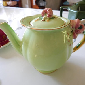 May include: A light green ceramic teapot with a matching lid, featuring a floral design and gold trim. The handle is adorned with pink and yellow accents. A burgundy teacup and saucer are visible in the background.