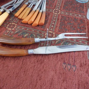 May include: A collection of vintage silverware with brown and cream-colored handles. The set includes a carving knife, carving fork, and various other utensils. The silverware is arranged on a patterned red tablecloth.