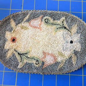 May include: Oval-shaped, hand-tufted rug with a floral design. The rug features a central cream-colored area with stylized flowers in shades of pink, beige, and green, surrounded by a gray border. The rug's edge is finished with a brown trim.