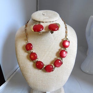 May include: A vintage jewelry set featuring a necklace and matching clip-on earrings. The necklace and earrings showcase large, round, red beads with a gold-tone metal chain and accents. The jewelry is displayed on a beige bust.