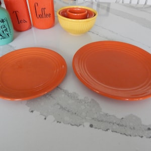 May include: Two orange ceramic dinner plates with a raised rim and a subtle ribbing pattern. The plates are on a white countertop.