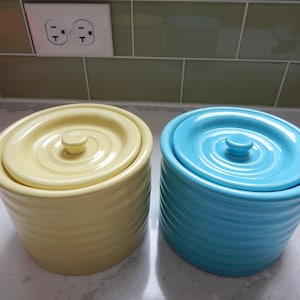 May include: Two ceramic canisters, one pale yellow and one turquoise blue, each with a matching lid and a small knob. The canisters have a ribbed texture and are suitable for kitchen storage.