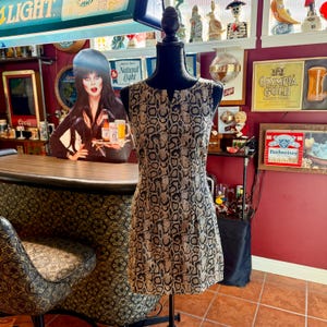 May include: A sleeveless dress with a snakeskin pattern in shades of brown and black is displayed on a mannequin. The dress has a fitted bodice and a slightly flared skirt. The background includes a bar and vintage decor.