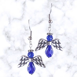 Pu&ograve; includere: Un paio di orecchini pendenti a forma di angelo in argento con accenti di cristallo blu. Gli angeli hanno ali d'argento e un corpo di cristallo blu.