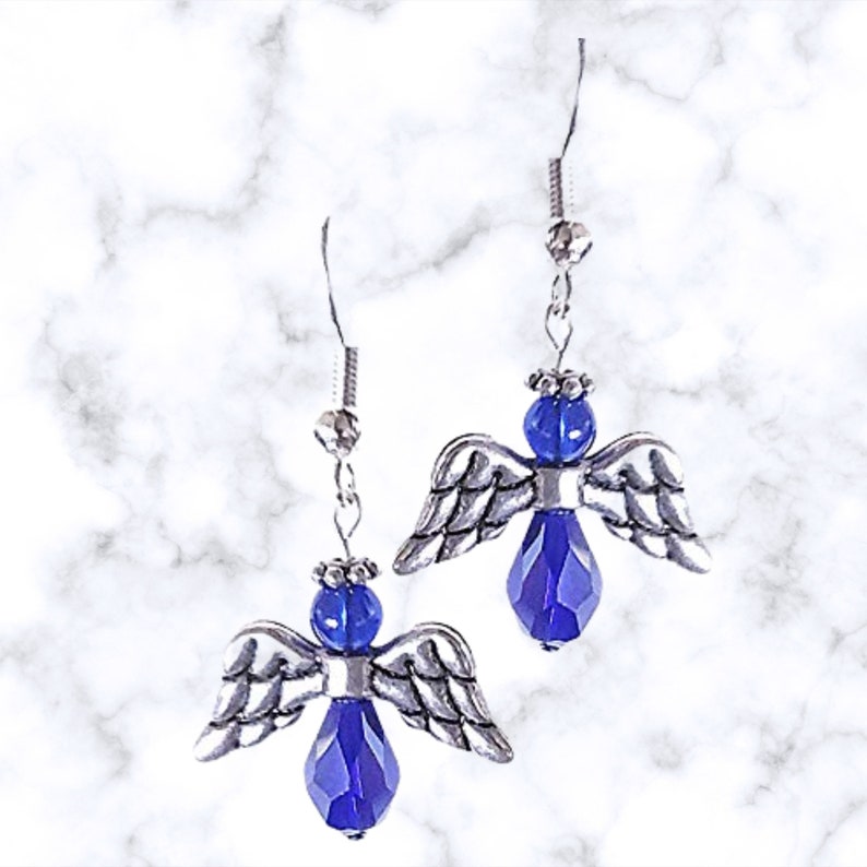 Pu&ograve; includere: Un paio di orecchini pendenti a forma di angelo in argento con accenti di cristallo blu. Gli angeli hanno ali d'argento e un corpo di cristallo blu.