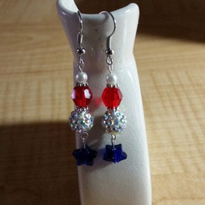 May include: A pair of dangle earrings with red, white, and blue beads. The earrings feature a star charm at the bottom.