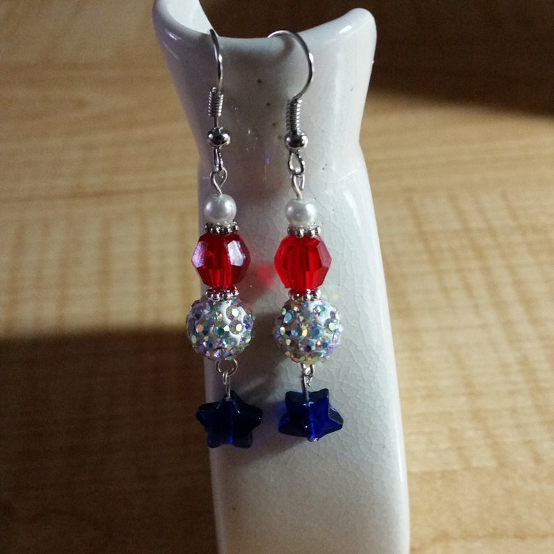 May include: A pair of dangle earrings with red, white, and blue beads. The earrings feature a star charm at the bottom.