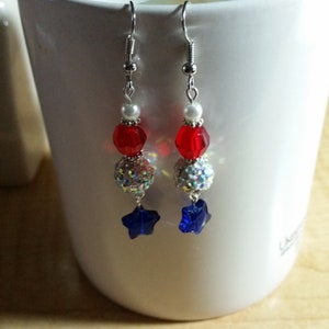 May include: Red, white, and blue star-shaped dangle earrings with silver hooks. The earrings feature a white pearl bead, a red crystal bead, a silver glitter ball, and a blue star-shaped crystal.