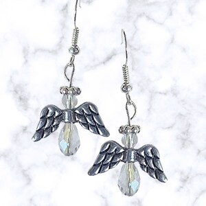 Pu&ograve; includere: Orecchini in argento con ali d'angelo e gocce di cristallo trasparente. Le ali sono dettagliate e i cristalli sono iridescenti.