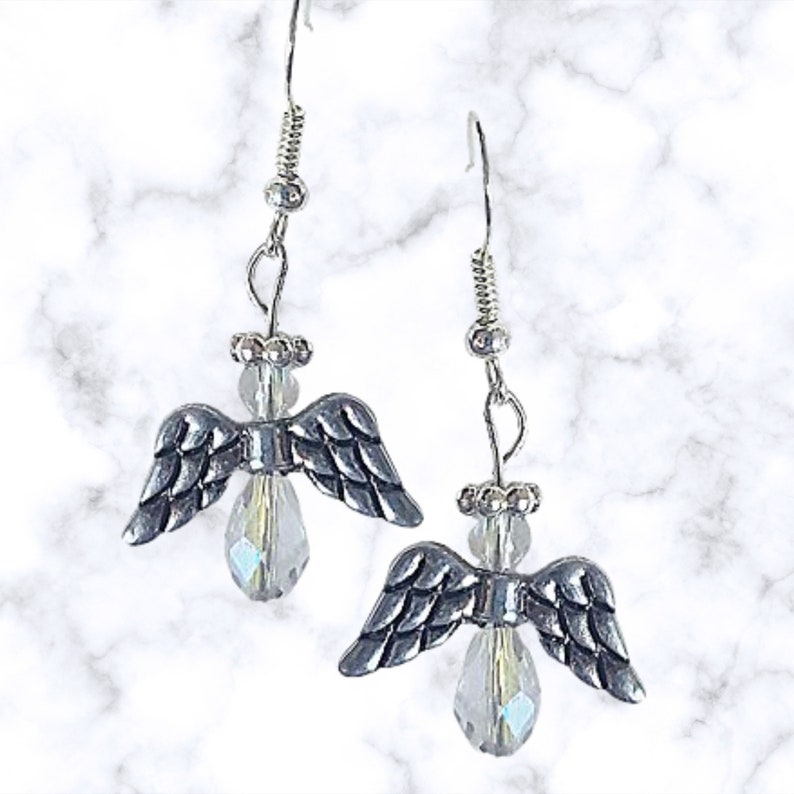 Pu&ograve; includere: Orecchini in argento con ali d'angelo e gocce di cristallo trasparente. Le ali sono dettagliate e i cristalli sono iridescenti.