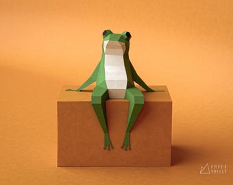 May include: A geometric paper frog in shades of green and white sits on a brown box against an orange background. The frog's body is made of folded paper, with its legs dangling over the edge. The Paper Valley logo is in the bottom right corner.
