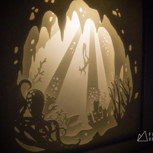 May include: A paper cut-out art piece depicting an underwater scene with a diver swimming through a cave, surrounded by coral and an octopus. The artwork is illuminated from behind, creating a glowing effect.