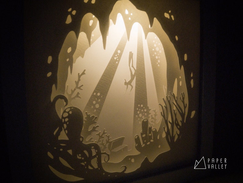 May include: A paper cut-out art piece depicting an underwater scene with a diver swimming through a cave, surrounded by coral and an octopus. The artwork is illuminated from behind, creating a glowing effect.