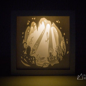 May include: A white paper light sculpture depicting an underwater scene with a diver swimming through a cave, surrounded by coral and an octopus. The light shines through the paper, creating a glowing effect.