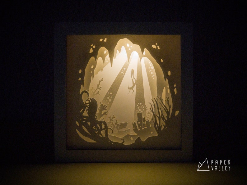 May include: A white paper light sculpture depicting an underwater scene with a diver swimming through a cave, surrounded by coral and an octopus. The light shines through the paper, creating a glowing effect.