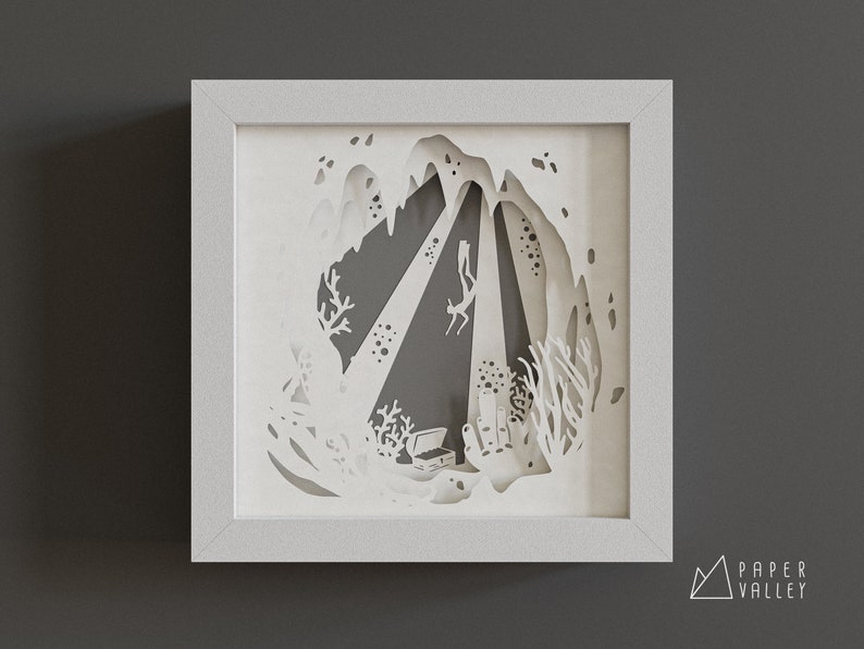 May include: White paper cut-out art depicting a diver exploring a cave with a treasure chest at the bottom. The art is framed in a white frame.