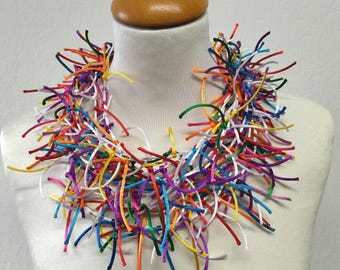 May include: A colorful necklace made of thin, knotted ribbons in various shades of red, orange, yellow, green, blue, purple, and white. The necklace is displayed on a white mannequin.