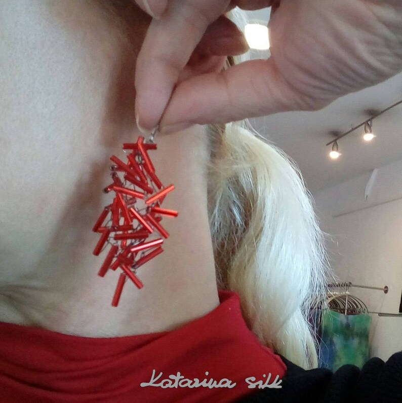 May include: Red beaded earrings with a silver hook. The earrings are made of small, rectangular beads that are strung together in a cascading design. The earrings are worn by a person with blonde hair.