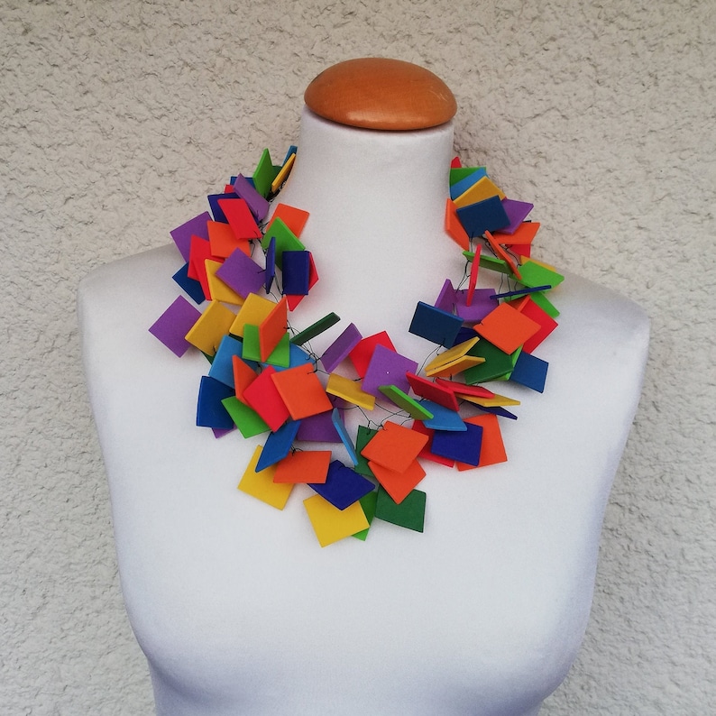 May include: A colorful necklace made of squares of felt in various colors, including red, orange, yellow, green, blue, purple, and pink. The necklace is displayed on a white mannequin.