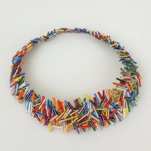 May include: A colorful necklace made of small, iridescent glass beads in various shades of red, orange, yellow, green, blue, and purple. The beads are arranged in a circular shape, creating a unique and eye-catching design.