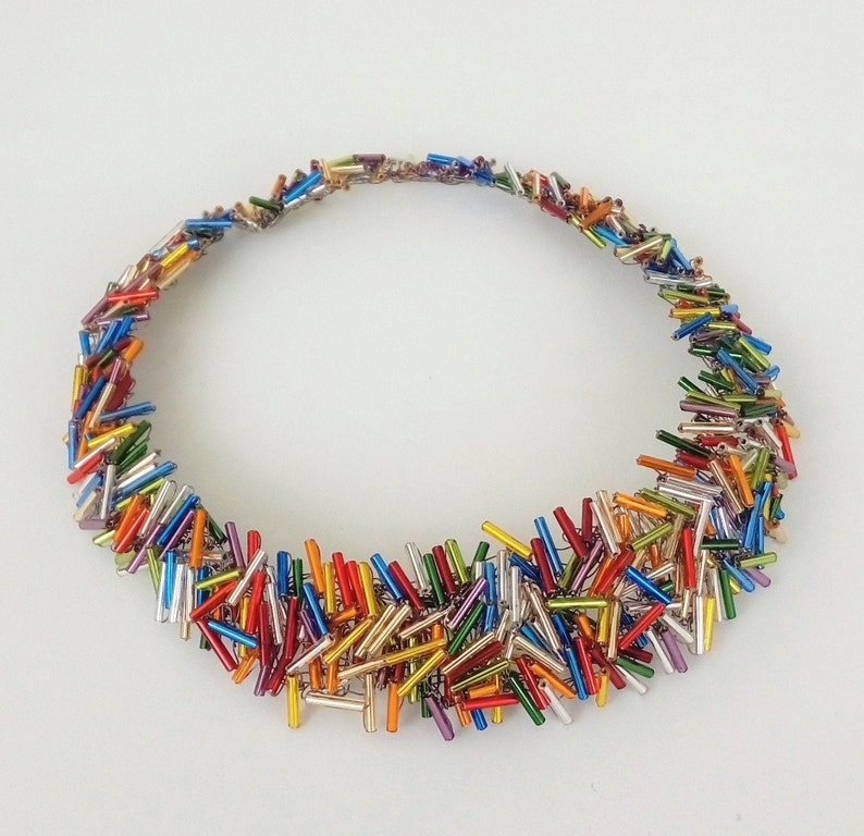 May include: A colorful necklace made of small, iridescent glass beads in various shades of red, orange, yellow, green, blue, and purple. The beads are arranged in a circular shape, creating a unique and eye-catching design.