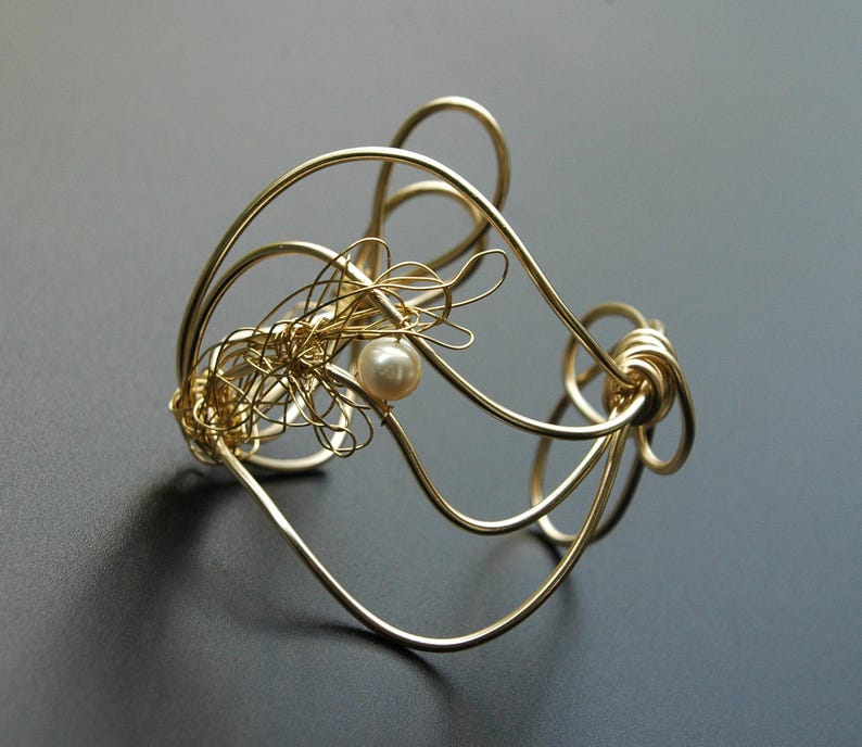 May include: A gold wire bracelet with a delicate pearl accent. The bracelet is made of multiple strands of wire that are intertwined to create a unique and intricate design.
