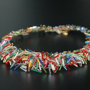 May include: A colorful necklace made of small, glass rods in red, blue, green, gold, and silver. The necklace is arranged in a circular shape, with the rods overlapping and creating a textured effect.