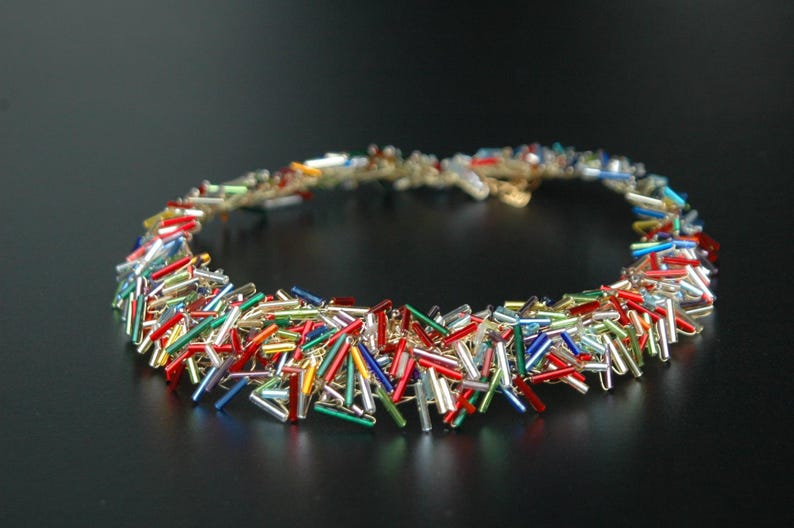 May include: A colorful necklace made of small, glass rods in red, blue, green, gold, and silver. The necklace is arranged in a circular shape, with the rods overlapping and creating a textured effect.