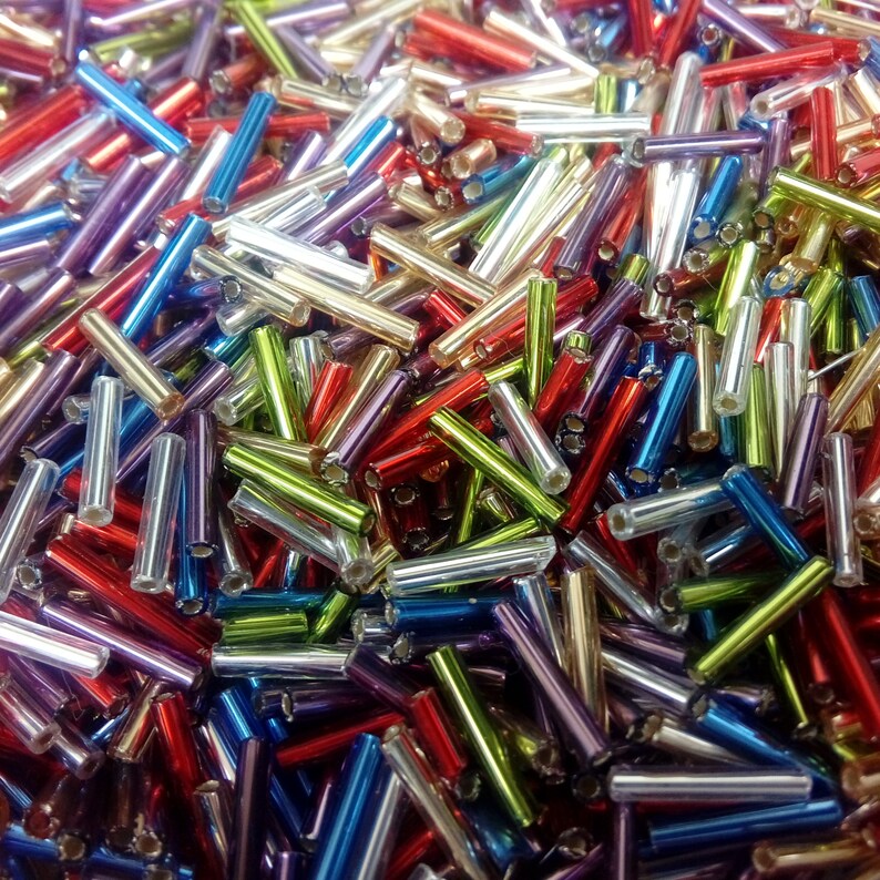 May include: A large assortment of colourful glass beads in various shades of red, blue, green, purple, and silver. The beads are all cylindrical in shape and have a shiny, metallic finish.