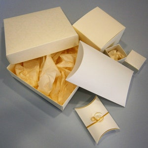 May include: A set of five ivory-colored gift boxes with tissue paper inside. The boxes are various sizes and shapes, including a rectangular box with a lid, a square box with a lid, and a pillow-shaped box.