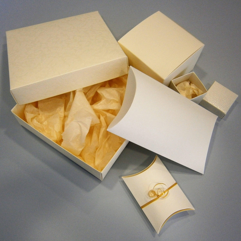 May include: A set of five ivory-colored gift boxes with tissue paper inside. The boxes are various sizes and shapes, including a rectangular box with a lid, a square box with a lid, and a pillow-shaped box.