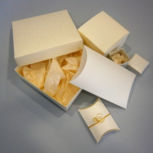 May include: A set of five ivory-colored gift boxes, including a larger box with a lid and tissue paper inside, a smaller box with a lid, and two pillow boxes. The pillow boxes are open and show a gold ribbon inside.