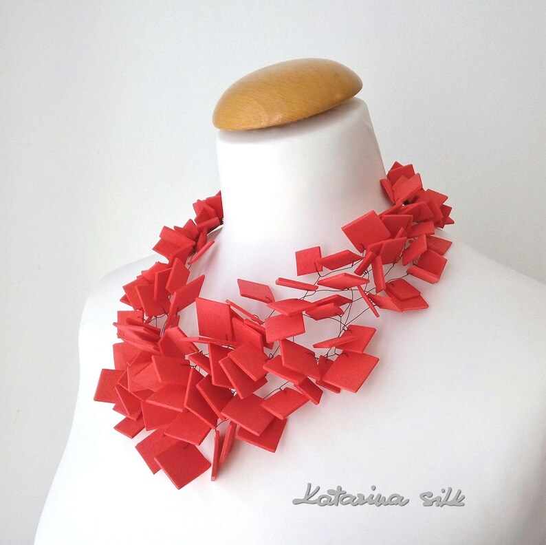 May include: A red statement necklace made of many small squares connected by thin wire. The necklace is displayed on a white mannequin bust.