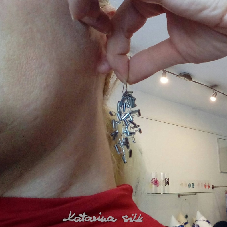 May include: A pair of silver earrings with a long, dangling design. The earrings are made of small, rectangular pieces of metal that are connected together. The earrings are hanging from a person's ear.
