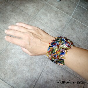 May include: A colorful bracelet made of small glass beads in various shades of red, orange, yellow, green, blue, and silver. The bracelet is worn on a person's wrist.