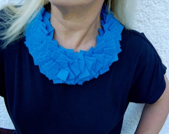 May include: A vibrant blue statement necklace composed of layered, square-cut fabric pieces. The necklace is worn over a dark blue top, creating a striking contrast. The design is a bold, geometric accessory.
