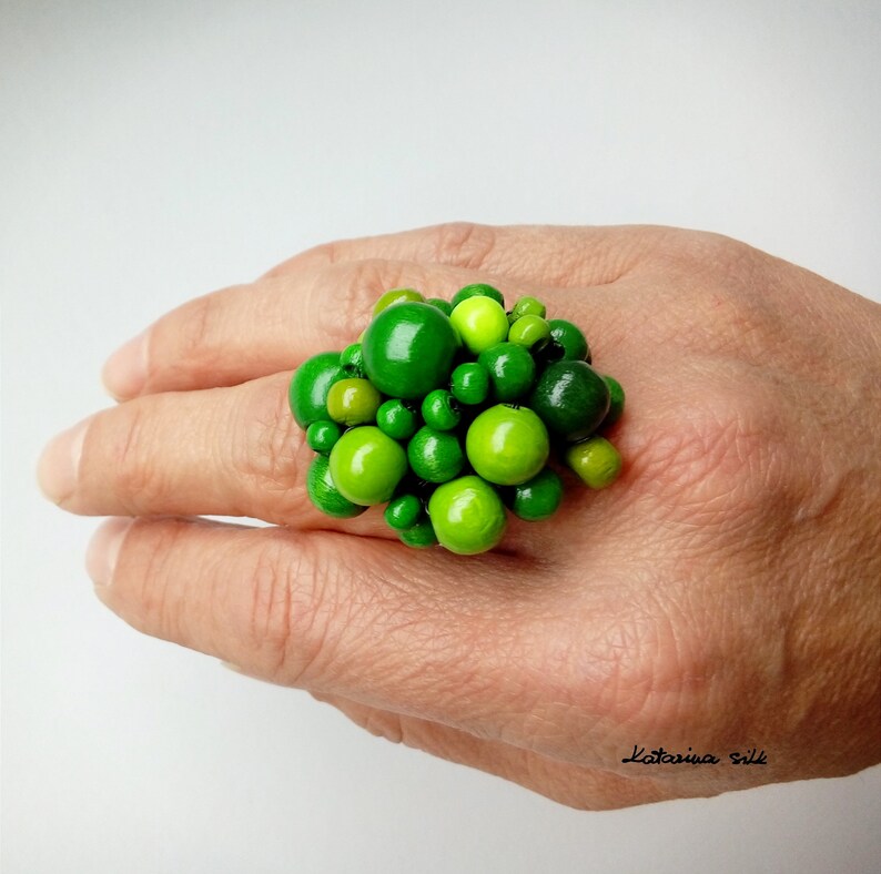 May include: A green statement ring made of many small green and lime green beads clustered together. The ring is worn on a person's finger.