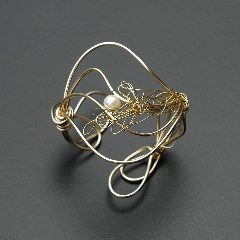 May include: A gold wire bracelet with a single pearl accent. The wire is twisted and formed into a delicate, organic shape.