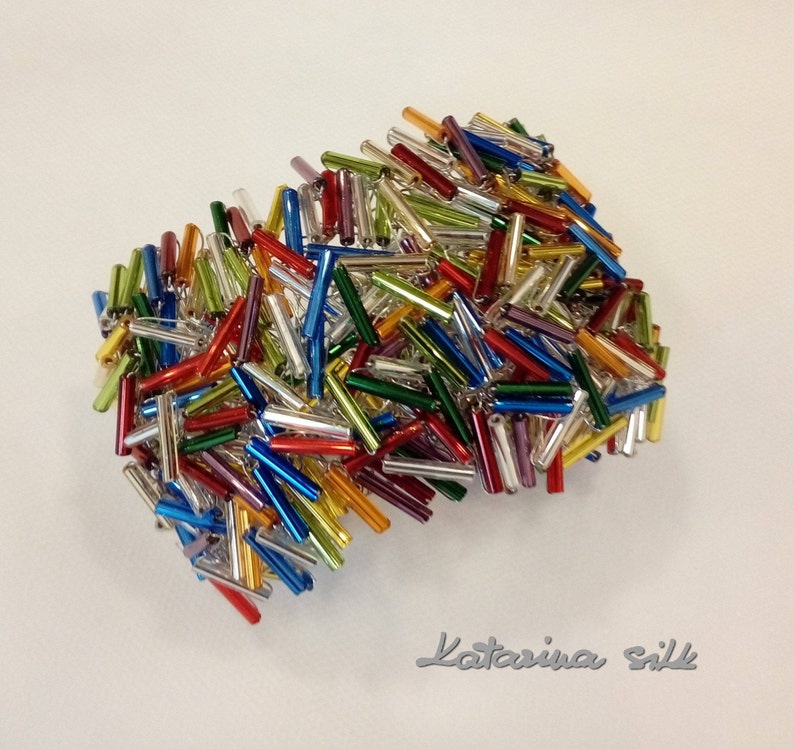 May include: A cluster of colorful glass beads in various shades of red, blue, green, yellow, and silver. The beads are arranged in a random pattern, creating a visually appealing and textured design.