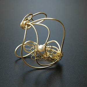 May include: A gold wire bracelet with a delicate pearl accent. The bracelet is made of multiple loops and swirls of wire, creating a unique and intricate design.