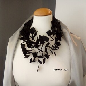 May include: A black necklace made of interconnected square shapes, worn on a white mannequin with a gray scarf. The necklace is made of a material that appears to be felt or fabric. The text "Katarina Silk" is visible in the bottom right corner of the image.