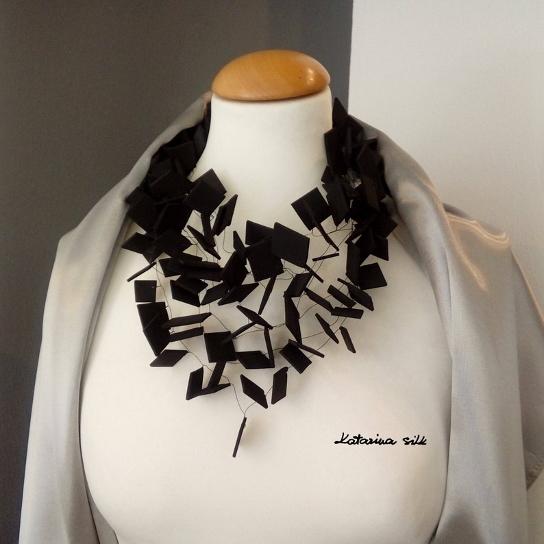 May include: A black necklace made of interconnected square shapes, worn on a white mannequin with a gray scarf. The necklace is made of a material that appears to be felt or fabric. The text "Katarina Silk" is visible in the bottom right corner of the image.
