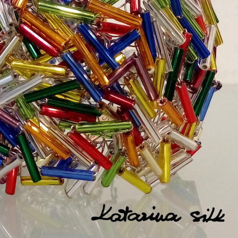 May include: A close-up of a colorful glass bead garland, featuring a mix of red, orange, yellow, green, blue, and silver beads. The beads are arranged in a random pattern, creating a visually appealing and textured design. The garland is perfect for adding a touch of color and whimsy to any space.