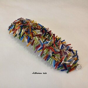 May include: A colorful bracelet made of small glass beads in various colors, including red, blue, green, yellow, and silver. The bracelet is made of a single strand of beads and has a silver clasp.